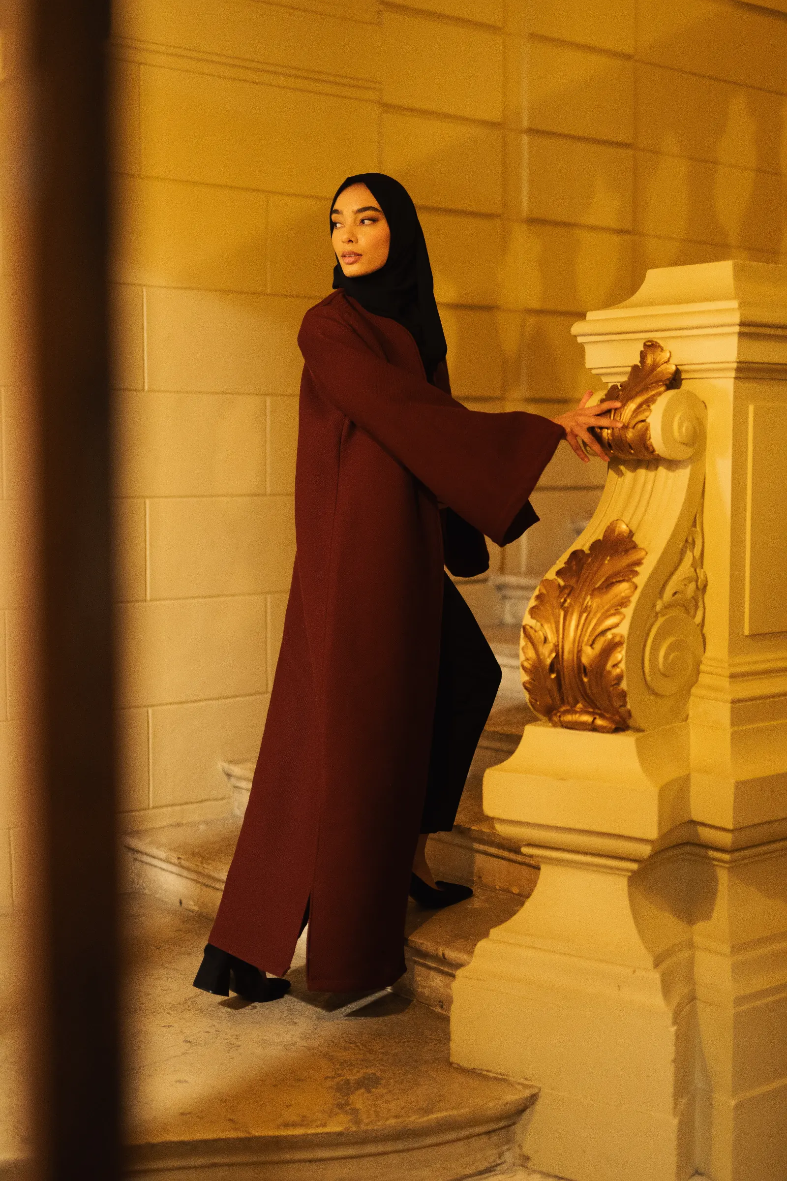 Femme en hijab noir et abaya bordeaux montant un escalier ancien avec ornements dorés.