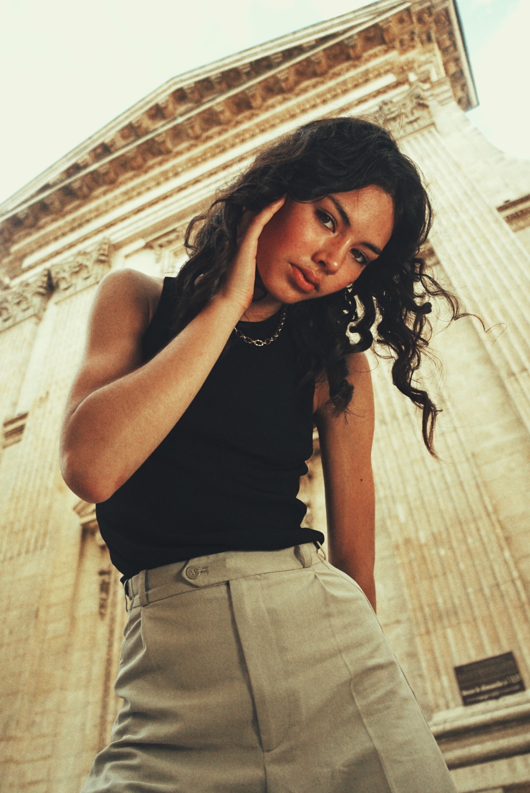 Femme aux cheveux bouclés, tee-shirt noir, pantalon clair, devant un bâtiment ancien.