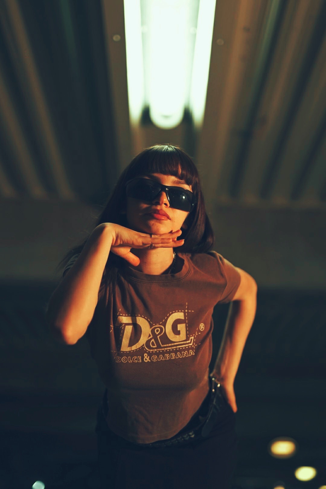 Femme avec frange, lunettes de soleil, t-shirt marron posant sous un néon lumineux.