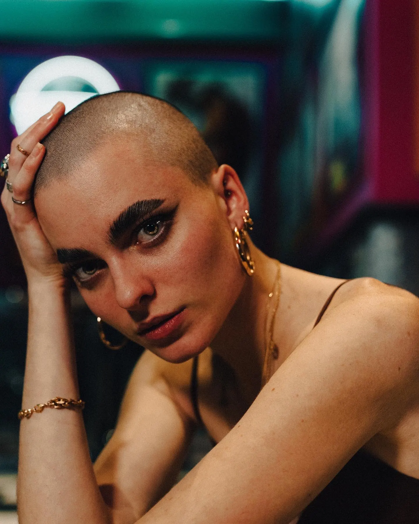 Personne au crâne rasé, boucles d'oreilles créoles dorées, maquillage intense des yeux.